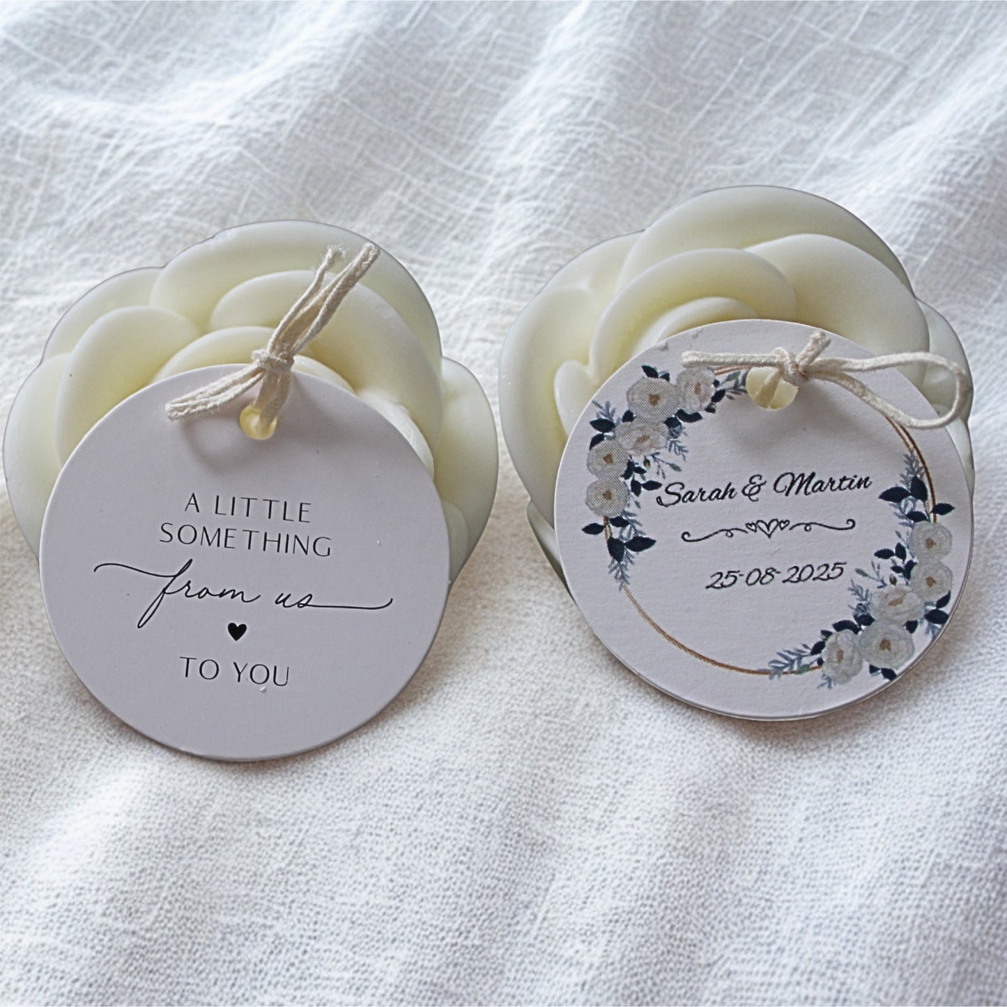 Two white wedding favour candles with personalised labels on white linen