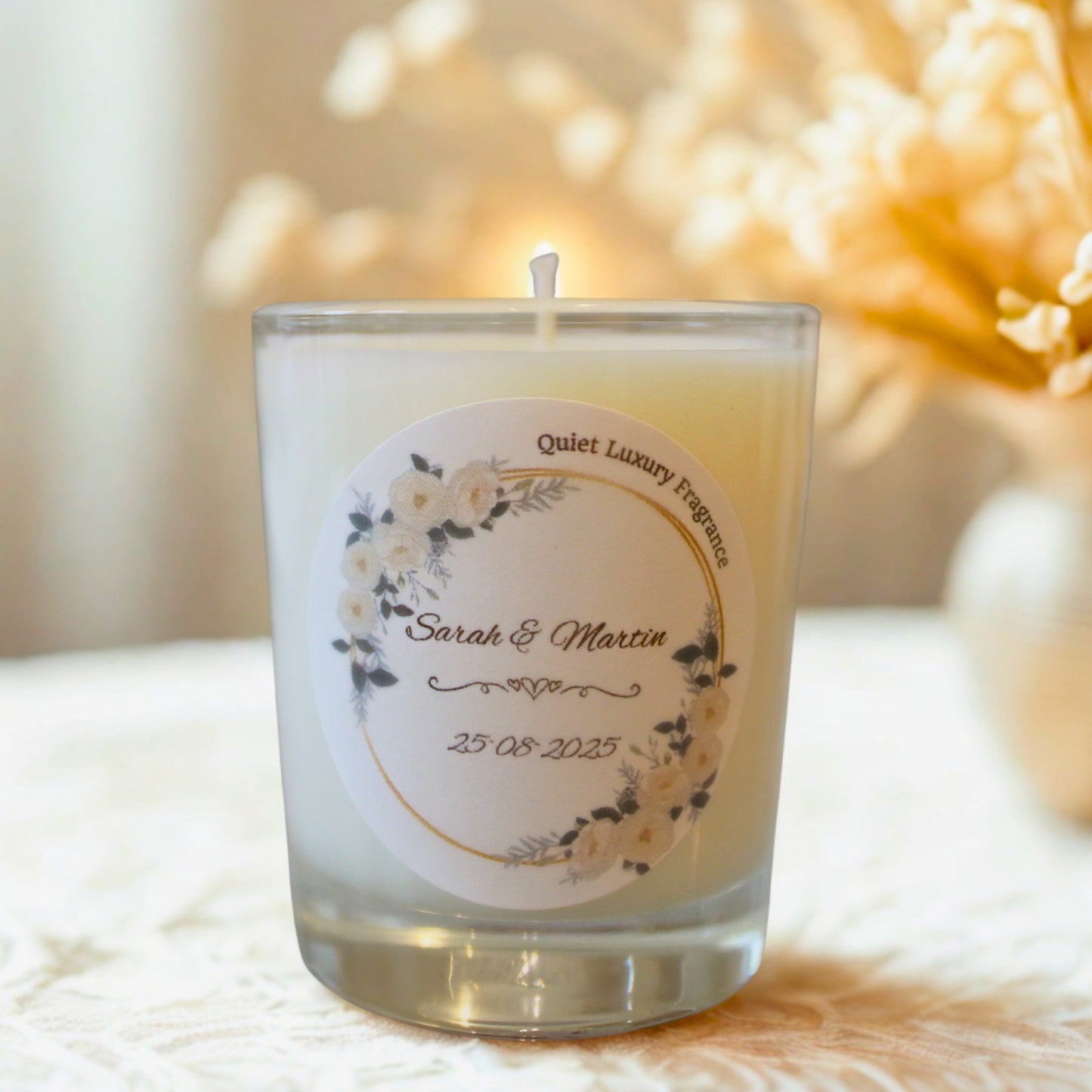 Personalized candle with a label featuring floral design and text, placed on a textured surface with a blurred decorative background.