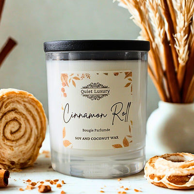 Candle labeled 'Cinnamon Roll' from Quiet Luxury with cinnamon roll and cinnamon sticks in the background.