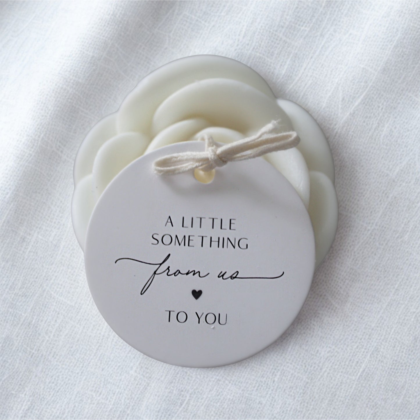 Round white soy wax favour with personalised gift tag with 'A little something from us to you' text on a white fabric background