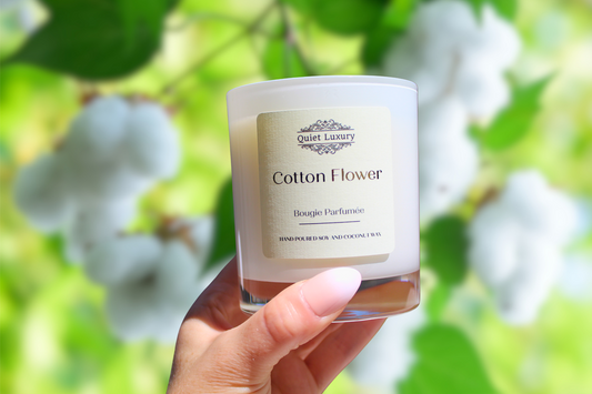 Candle labeled 'Cotton Flower' held in front of a blurred natural background with greenery and white flowers.