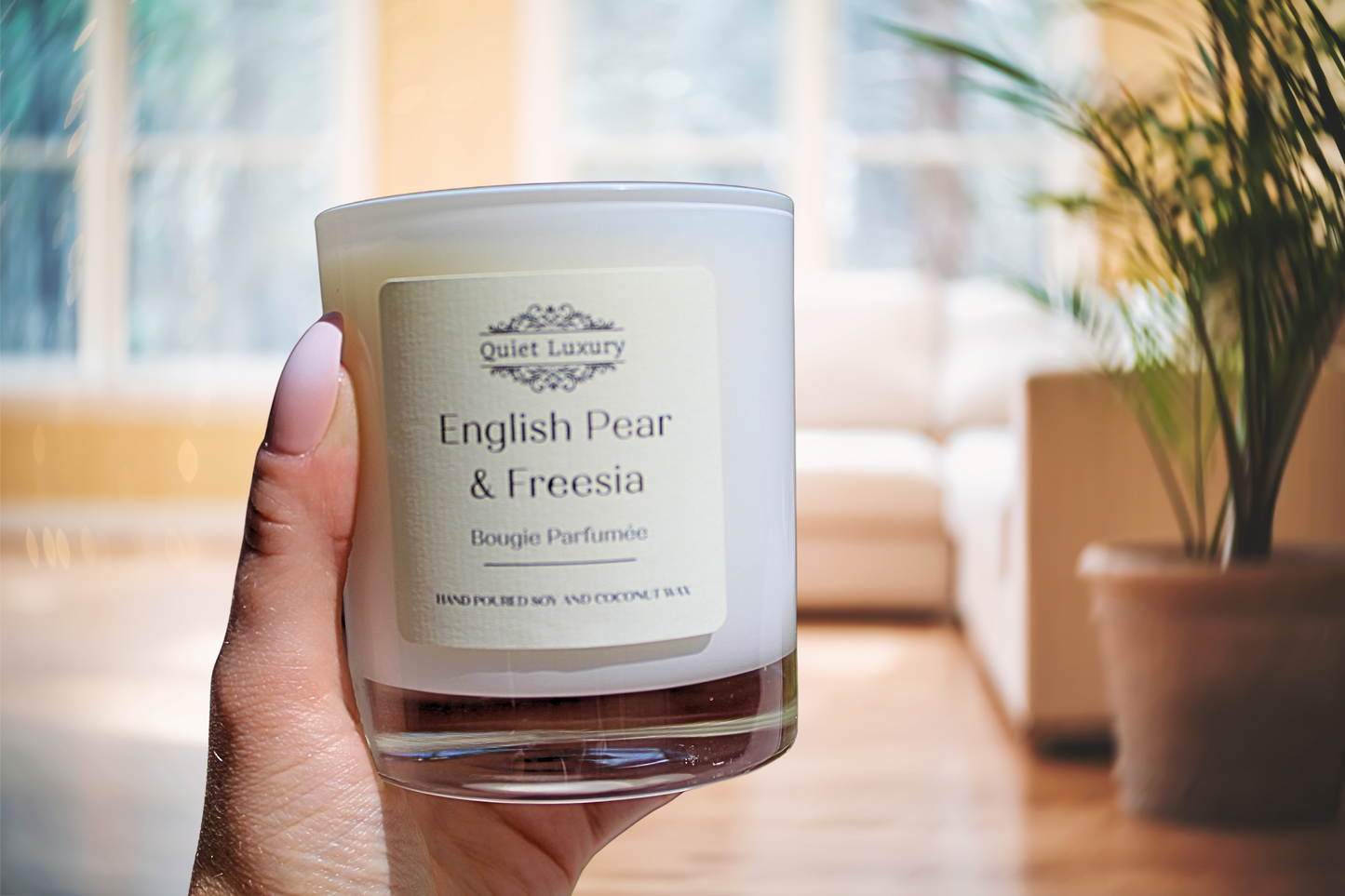 Hand holding a 'Quiet Luxury English Pear & Freesia' candle in a room with a plant and window.