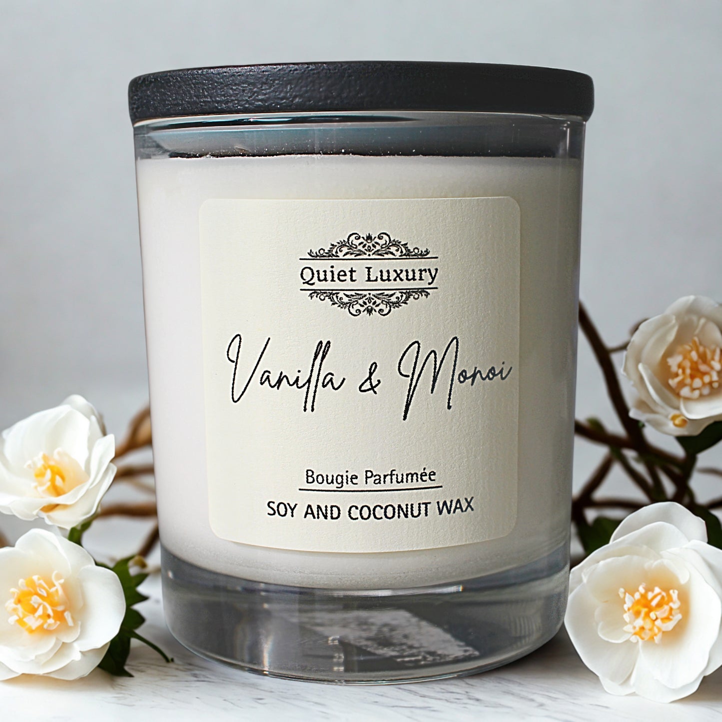 Candle labeled 'Quiet Luxury Vanilla & Monoi' with white flowers in the background